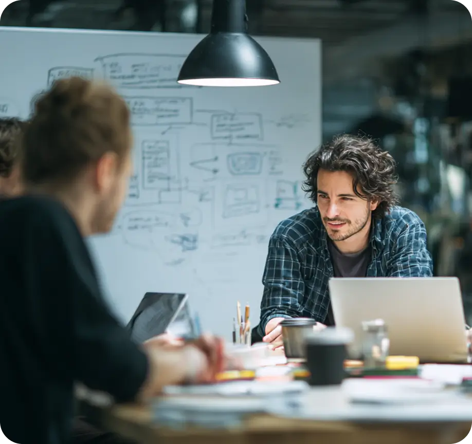 Dos personas del equipo de Koombea trabajando una idea de producto en una mesa, con un tablero de flujos detrás