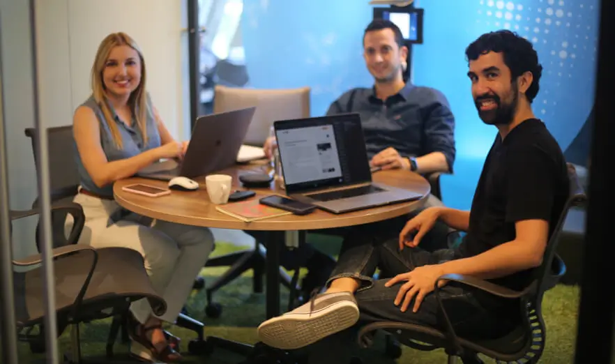 Tres personas del equipo de Koombea en una mesa redonda con portátiles abiertos, mirando a la cámara